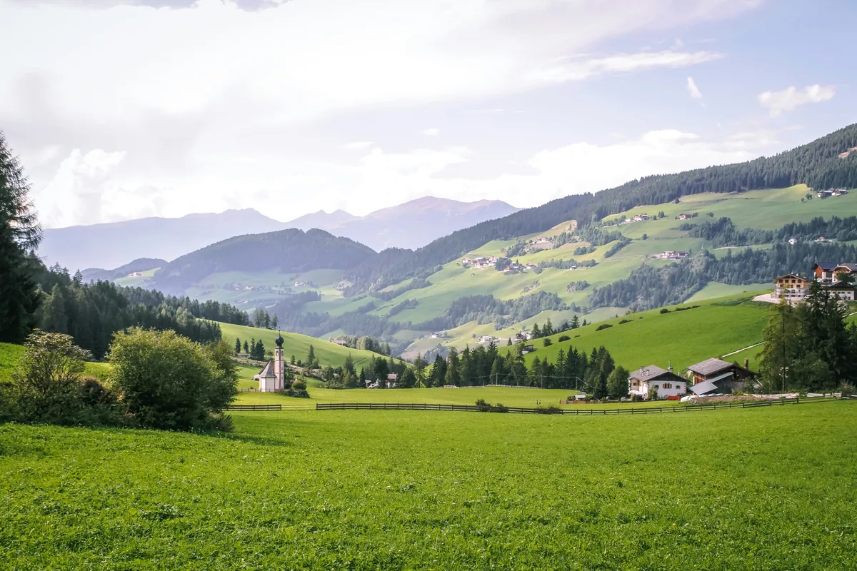 Grüne Almlandschaft mit Kapelle, Bauernhöfen, Wäldern und Bergen